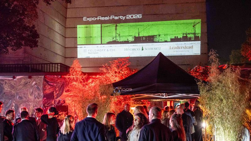 Eine abendliche Szene bei der Expo-Real-Party 2026, bei der eine Projektion mit Firmenlogos und Baustellen-Grafiken auf die Fassade eines Gebäudes geworfen wird, während Gäste in einer festlich beleuchteten Außenanlage mit rotem Licht und Bambusdekor feiern.