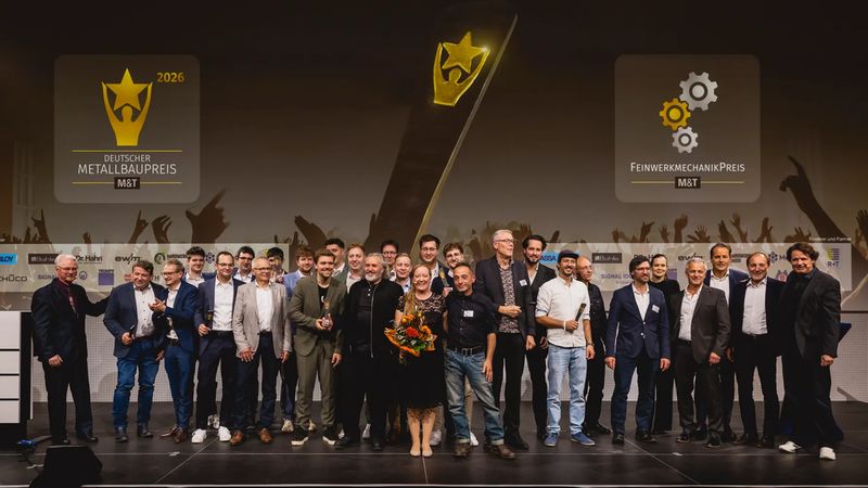Eine große Gruppe von Preisträgern und Verantwortlichen posiert auf einer Bühne nach der Verleihung des Deutschen Metallbaupreises und des Feinwerkmechanikpreises 2026. Im Hintergrund sind die entsprechenden Logos auf großen Bildschirmen zu sehen, während die Gruppe freudig in die Kamera lächelt.