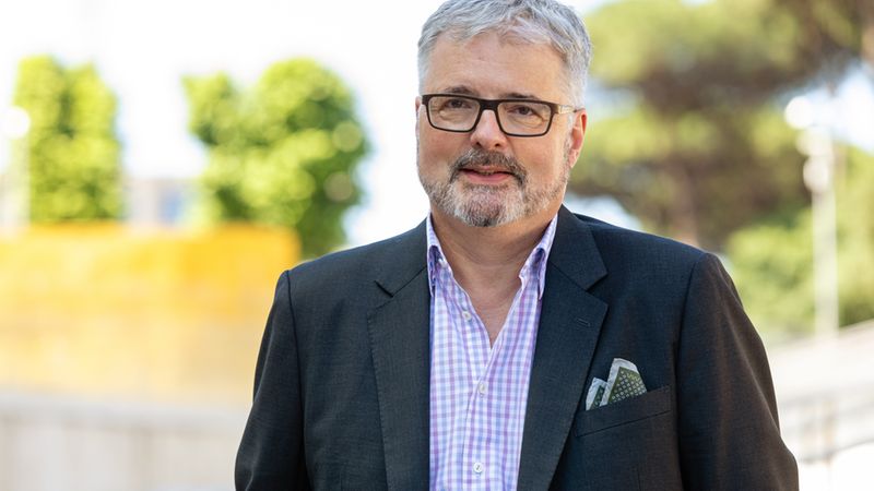 Ein Mann mit grauen Haaren und Brille trägt einen Anzug und steht im Freien vor einem unscharfen Hintergrund mit Bäumen.
