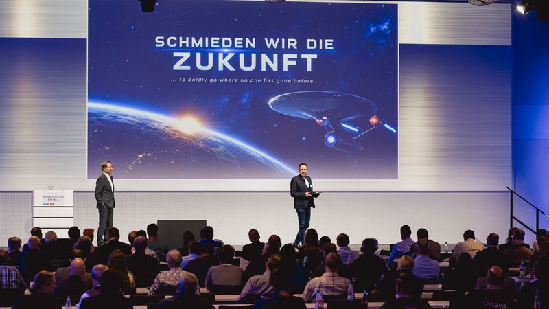 Ein Redner steht auf einer Bühne vor einem großen Bildschirm, auf dem der Slogan „Schmieden wir die Zukunft“ und ein Raumschiff vor einer Erde bei Sonnenaufgang zu sehen sind, während ein Publikum die Präsentation verfolgt.