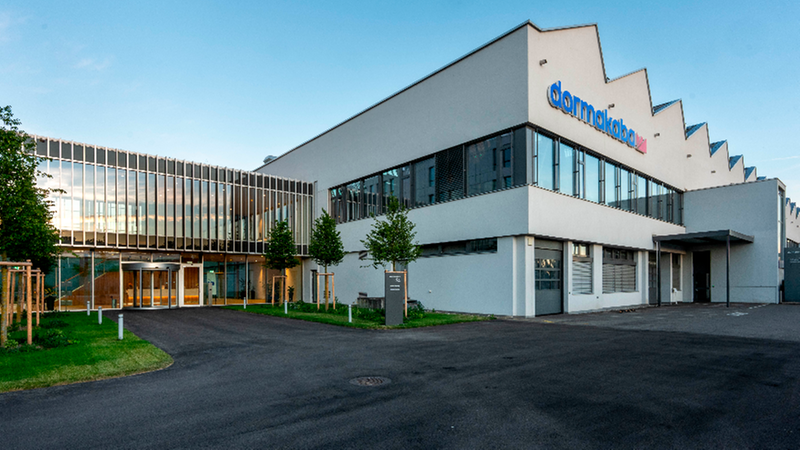 Eine Außenansicht des Firmengebäudes von dormakaba bei Tageslicht mit modernem Architekturstil, Glasfassade und begrünten Außenanlagen.