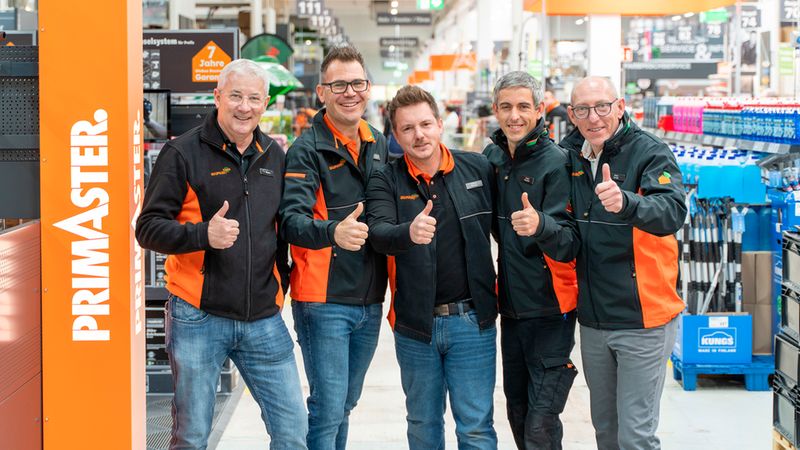 Ein freundliches Team von fünf Mitarbeitern des Primaster-Baumarkts posiert gemeinsam in den Gängen des Geschäfts und zeigt enthusiastisch den Daumen nach oben.