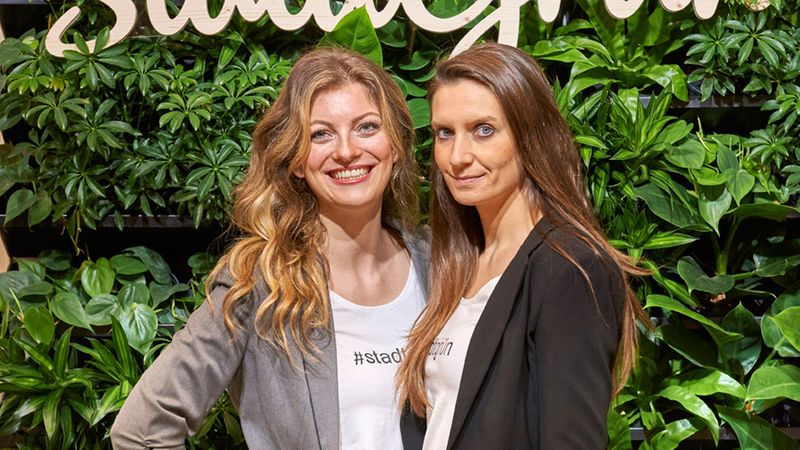 Zwei Frauen stehen vor einer grünen Wand mit dem Logo "StadtGrün" auf einer Messe.