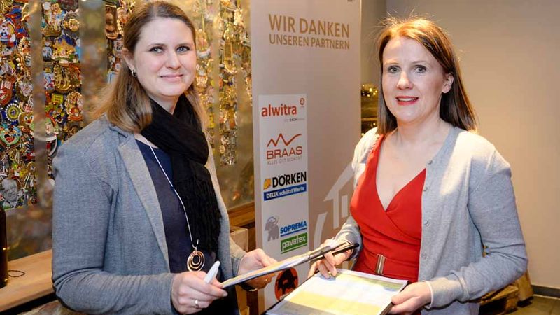 Zwei Frauen stehen bei einem Event vor einem Banner mit Sponsorenlogos und halten Klemmbretter in den Händen.
