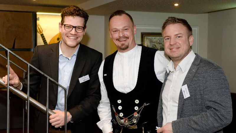 Eine Gruppe von drei Männern in formeller Kleidung, die in einem Innenraum lächeln.