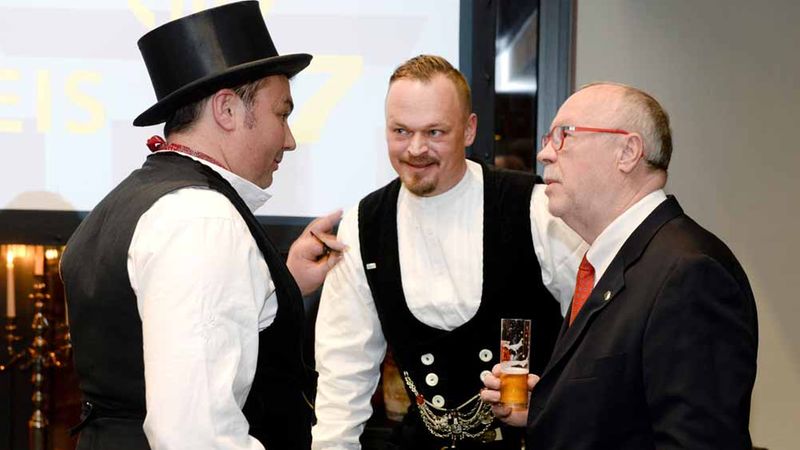 Drei Männer in traditioneller Kleidung unterhalten sich in einem Raum. Einer der Männer hält ein Glas Bier in der Hand.