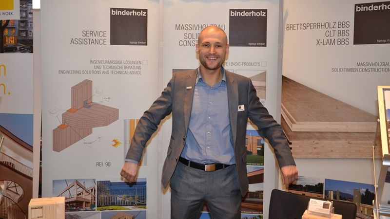 Ein Messeaussteller von binderholz steht lächelnd vor einem Informationsstand mit verschiedenen Holzprodukten.