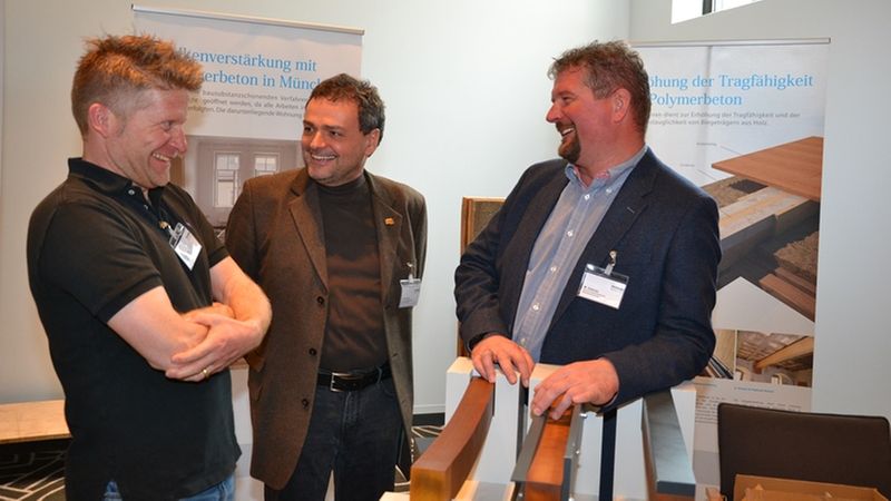 Drei Männer unterhalten sich angeregt auf einer Messe über Bauprodukte.