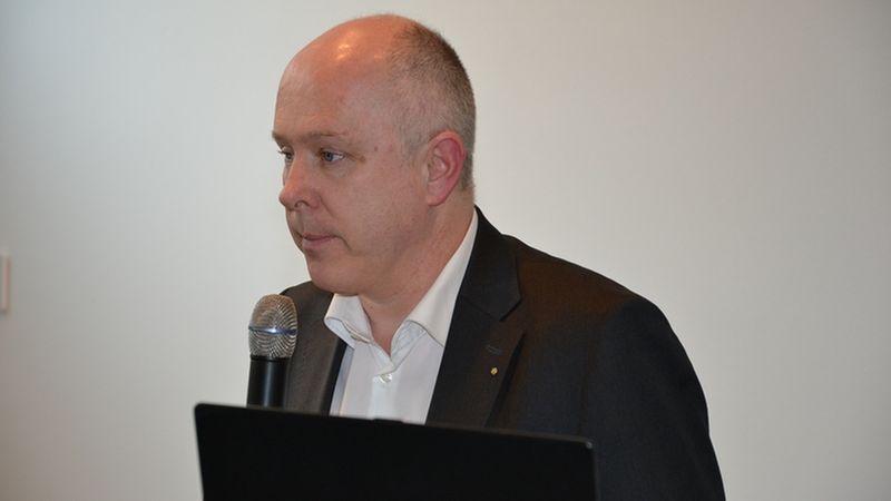 Ein Mann in einem Anzug hält ein Mikrofon und spricht vor einem Publikum.
