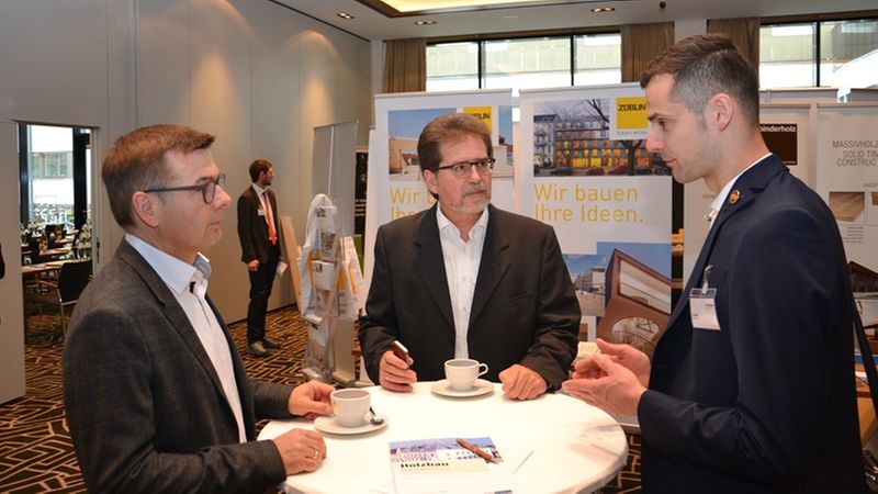 Drei Männer in Anzügen diskutieren an einem Kaffeetisch während einer Konferenz.