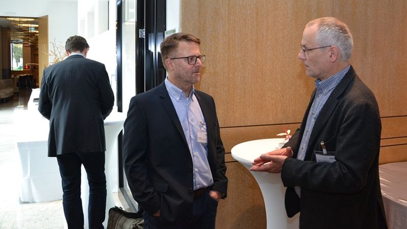 Zwei Männer im Anzug stehen an einem Stehtisch und unterhalten sich auf einer Konferenz.