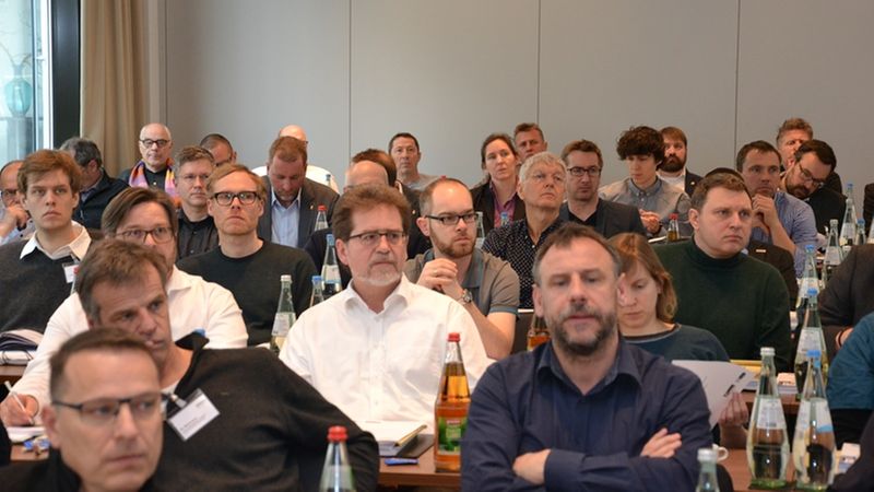 Eine Gruppe von Menschen sitzt aufmerksam in einem Konferenzraum und hört einem Vortrag zu.