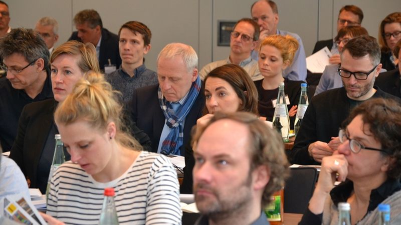 Eine Gruppe von Menschen sitzt aufmerksam in einem Konferenzraum und hört einem Vortrag zu.