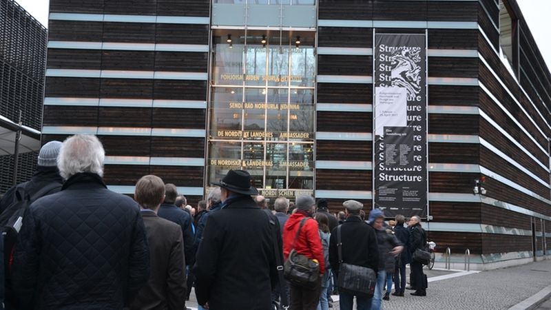 Eine Gruppe von Menschen steht vor einem modernen Gebäude mit einer auffälligen Glasfassade und einem großen Banner an der Seite.