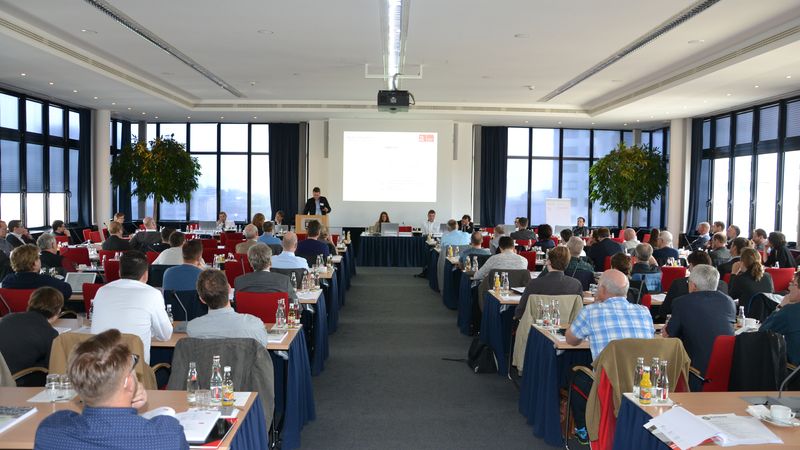 Ein großer Konferenzraum mit vielen Teilnehmern, die einer Präsentation folgen.