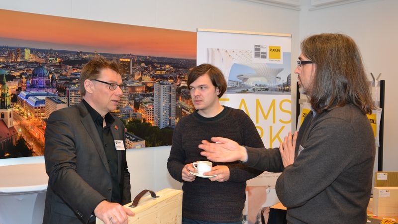 Drei Männer im Gespräch an einem Messestand mit einem großen Stadtbild im Hintergrund.