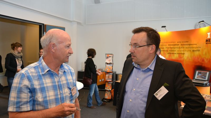 Zwei Männer führen ein Gespräch auf einer Messe, umgeben von Informationsständen und anderen Teilnehmern.