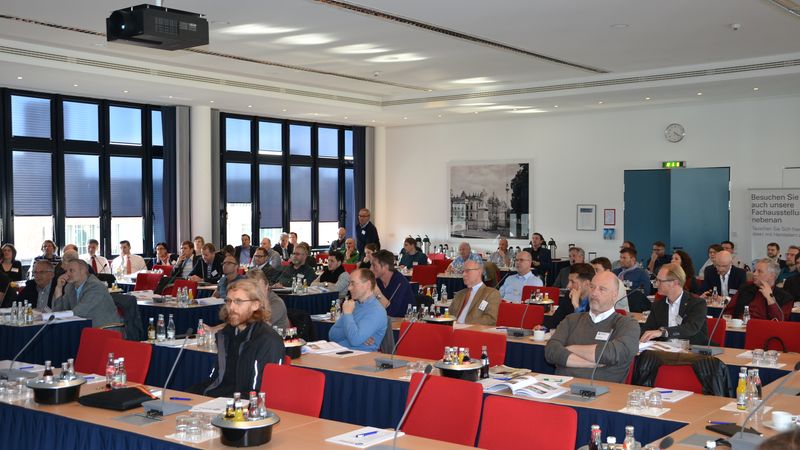 Ein Konferenzraum voller Teilnehmer, die einer Präsentation folgen. Die Teilnehmer sitzen an Tischen mit Getränken und Unterlagen.