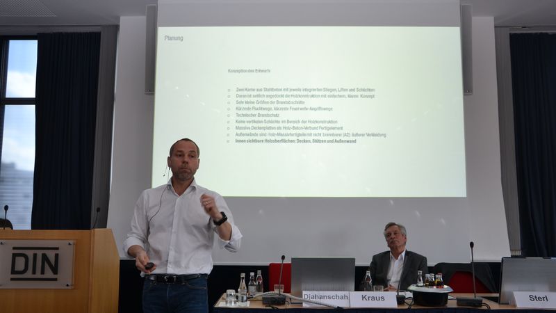 Ein Mann hält einen Vortrag bei einer Konferenz, während ein anderer Mann am Tisch sitzt. Im Hintergrund ist eine Präsentation zu sehen.