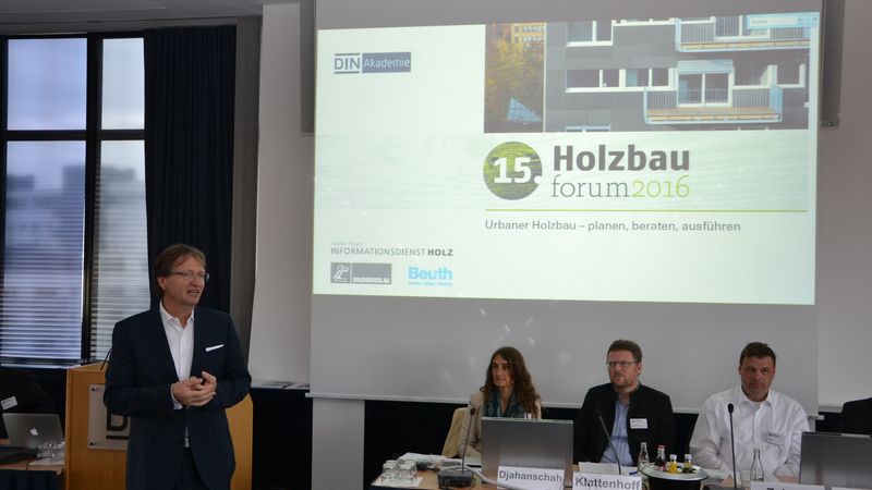 Ein Redner spricht auf dem Holzbau Forum 2016 vor einer Projektionsleinwand, während vier Personen an einem Tisch sitzen.