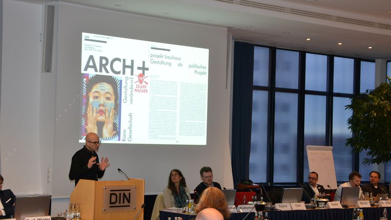 Ein Redner hält eine Präsentation bei einer Konferenz im DIN-Gebäude, während das Publikum aufmerksam zuhört.