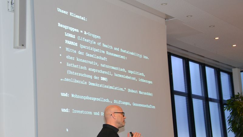 Ein Mann hält einen Vortrag vor einer Projektionsfläche mit Text über Baugruppen und Konsumenten.