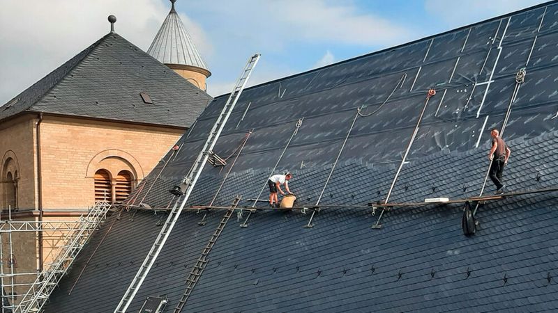 Zwei Dachdecker arbeiten auf einem großen Schieferdach eines Gebäudes mit Türmen im Hintergrund.