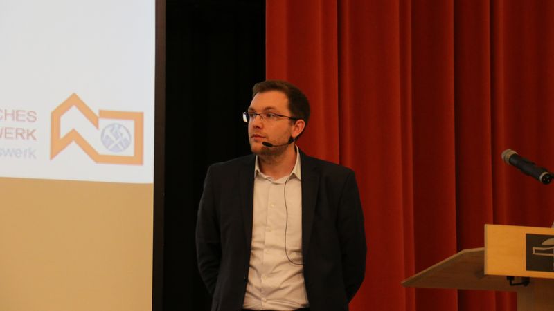 Ein Mann hält einen Vortrag auf einer Bühne mit rotem Vorhang im Hintergrund.