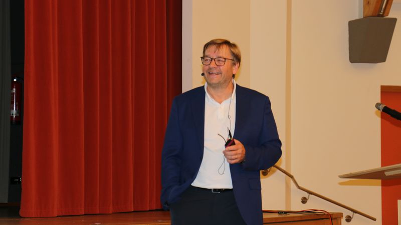 Ein Mann in einem blauen Anzug hält einen Vortrag auf einer Bühne mit rotem Vorhang im Hintergrund.