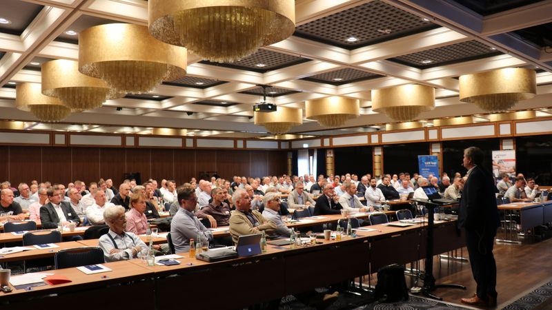 Ein großer Konferenzraum voller Teilnehmer, die einem Vortrag lauschen.