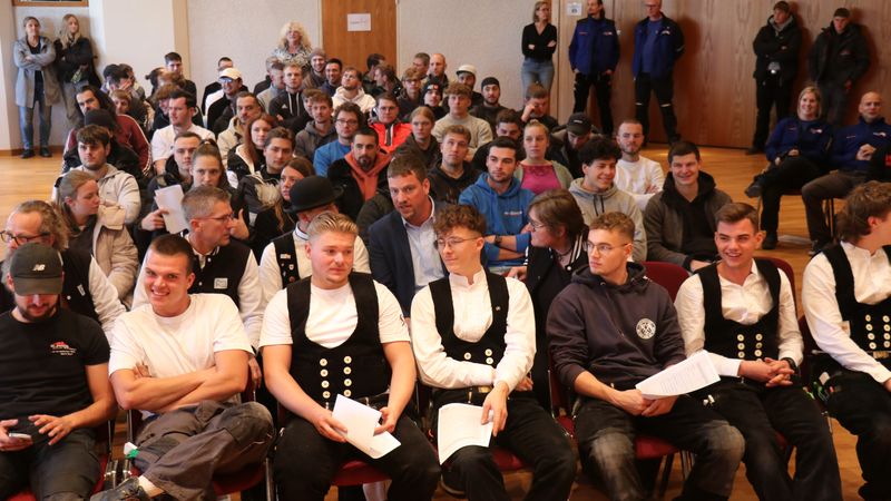 Eine Gruppe von Handwerkern in traditioneller Kleidung sitzt in einem Versammlungsraum.