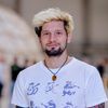 Ein freundliches Porträt eines jungen Mannes mit markanter blond-schwarzer Frisur, einem gepflegten Bart und einem weißen T-Shirt mit Musik machenden Tier-Illustrationen, der einen auffälligen Anhänger an einer schwarzen Kette trägt.