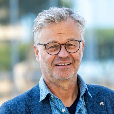 Ein Mann mit Brille und grauen Haaren lächelt in die Kamera. Er trägt ein blaues Sakko und ein gemustertes Hemd.