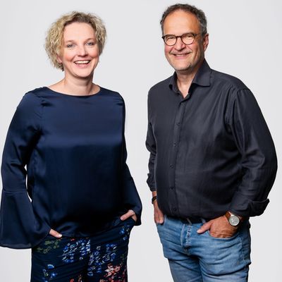 Ein freundliches, professionelles Porträt eines Unternehmerpaares, das gemeinsam vor einem neutralen, hellgrauen Hintergrund steht und in die Kamera lächelt.
