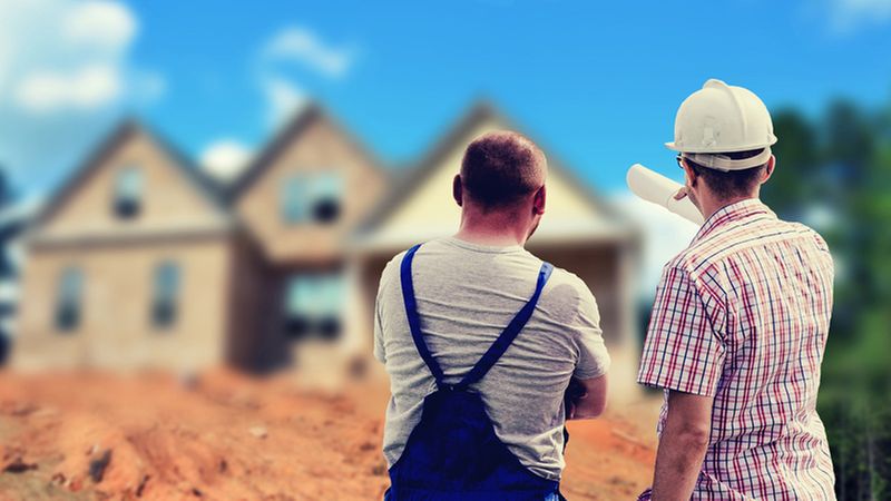 Ein Mann in blauer Arbeitskleidung und ein Mann im karierten Hemd mit weißem Bauhelm schauen auf eine Baustelle.