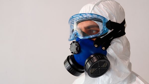 Hauptmotiv: Eine Person trägt einen weißen Schutzanzug, eine blaue Filter-Atemschutzmaske und eine transparente Schutzbrille. Die Nahaufnahme zeigt die Filterpatronen, Befestigungsriemen und die Abdichtung der Maske vor einem neutralen Hintergrund. Geeignet zur Darstellung von persönlicher Schutzausrüstung, Arbeitssicherheit, Labor- oder Gefahrstoffschutz.