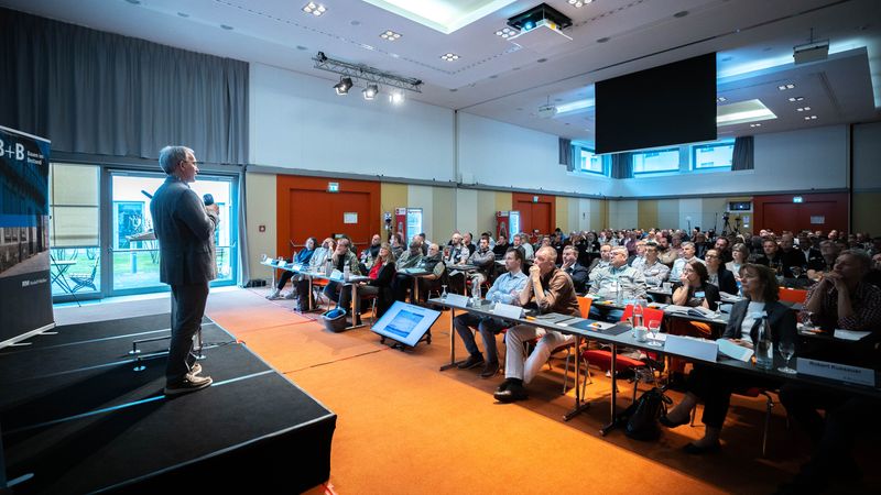 Ein Redner hält einen Vortrag vor einem großen Publikum in einem Konferenzraum.