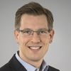 Ein Mann mit Brille lächelt in die Kamera. Er trägt ein Sakko und ein kariertes Hemd.