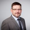 Ein Mann mit Brille und Bart in einem grauen Anzug und Krawatte lächelt in die Kamera.