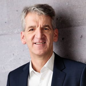 Ein Mann in einem dunklen Anzug steht vor einer grauen Betonwand und lächelt in die Kamera.