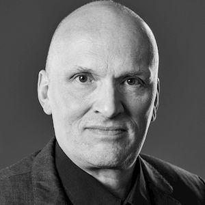 Ein Schwarz-Weiß-Porträt eines Mannes mit ernstem Gesichtsausdruck vor einem dunklen Hintergrund.