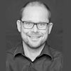 Dipl.-Ing. Kai Schulz. Ein schwarz-weiß Porträt eines lächelnden Mannes mit Brille vor einem dunklen Hintergrund.