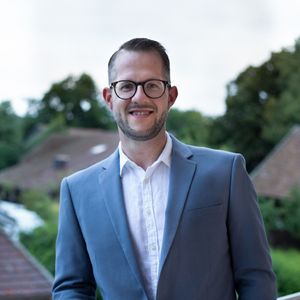 Ein Mann im Anzug steht auf einem Balkon mit einem Lächeln im Gesicht.