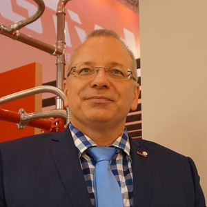 Ein Mann in einem blauen Anzug mit einer blauen Krawatte steht vor einer industriellen Kulisse mit Rohren.