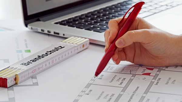 
    Ein Bild von einem Arbeitsplatz mit einem offenen Laptop und einem Brandschutzplan, auf dem mit einem roten Stift markiert wird. Neben dem Laptop liegt ein Zollstock. Der Fokus liegt auf den Händen der Person, die den Stift hält und auf dem Brandschutzplan arbeitet.
  