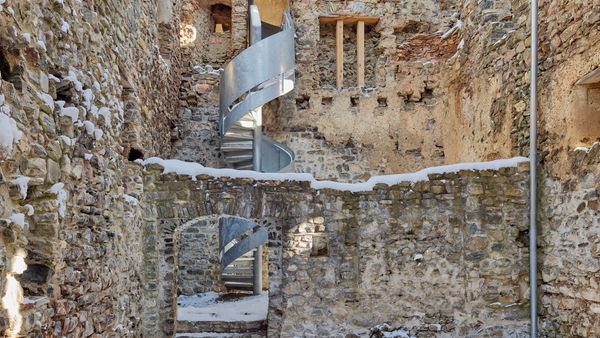Eine alte Burgruine mit einer modernen, schneebedeckten Wendeltreppe im Inneren. Die Mauern sind aus grobem Stein und teilweise mit Schnee bedeckt.