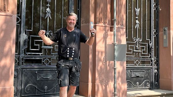 Ein Handwerker steht lächelnd vor einer kunstvoll verzierten Haustür und hält ein Werkzeug in der Hand.