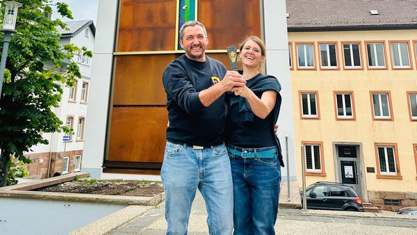 Zwei Personen stehen vor einem hohen Kirchturm und halten gemeinsam einen Pokal.