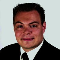 Ein Porträt eines lächelnden Mannes in Geschäftskleidung, mit Anzug und Krawatte, vor einem weißen Hintergrund.