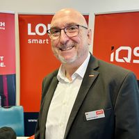 Ein lächelnder Mann mit Brille steht vor einem roten Banner auf einer Messe oder Veranstaltung.